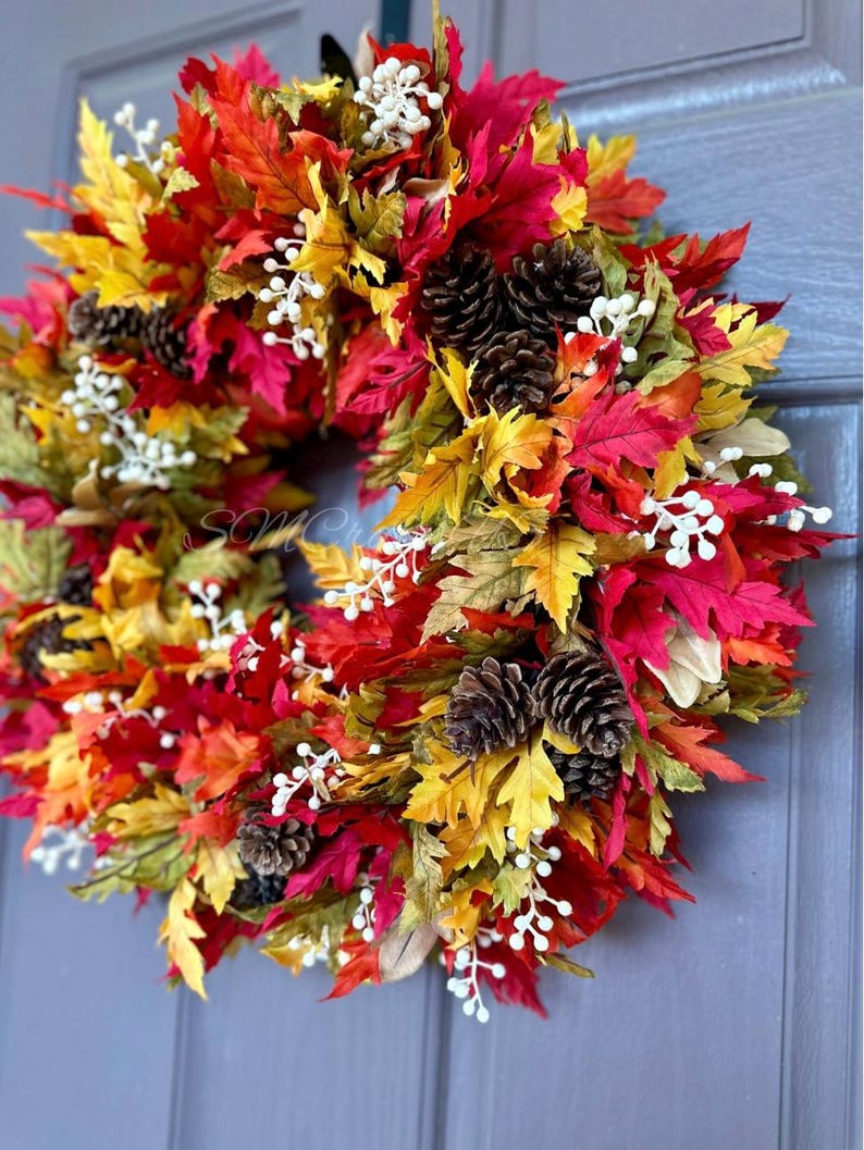 K&ouml;nnte beinhalten: Ein farbenfroher Herbstkranz mit roten, gelben und gr&uuml;nen Bl&auml;ttern. Der Kranz ist mit Tannenzapfen und kleinen wei&szlig;en Beeren geschm&uuml;ckt. Die saisonale Dekoration h&auml;ngt an einer blauen T&uuml;r.