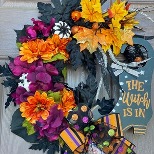 May include: A Halloween wreath with a witch's boot, black, orange, and purple flowers, and a black and orange striped bow with polka dots. The sign says "The Witch Is In".
