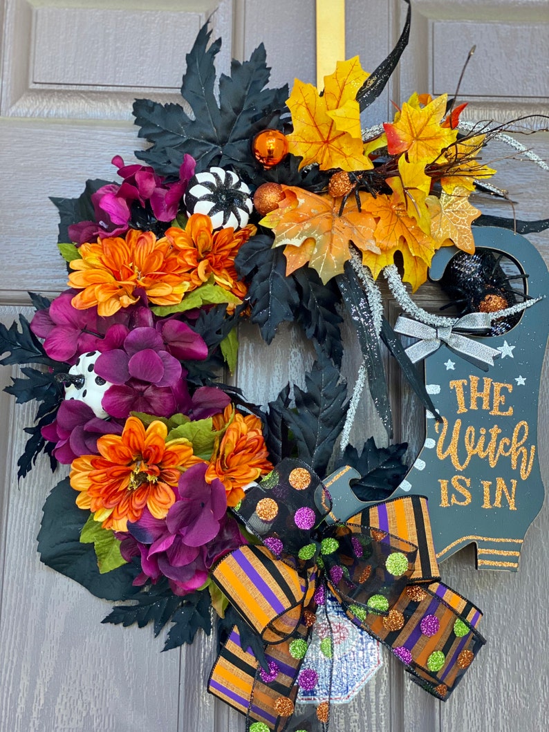 May include: A Halloween wreath with a witch's boot, black, orange, and purple flowers, and a black and orange striped bow with polka dots. The sign says "The Witch Is In".