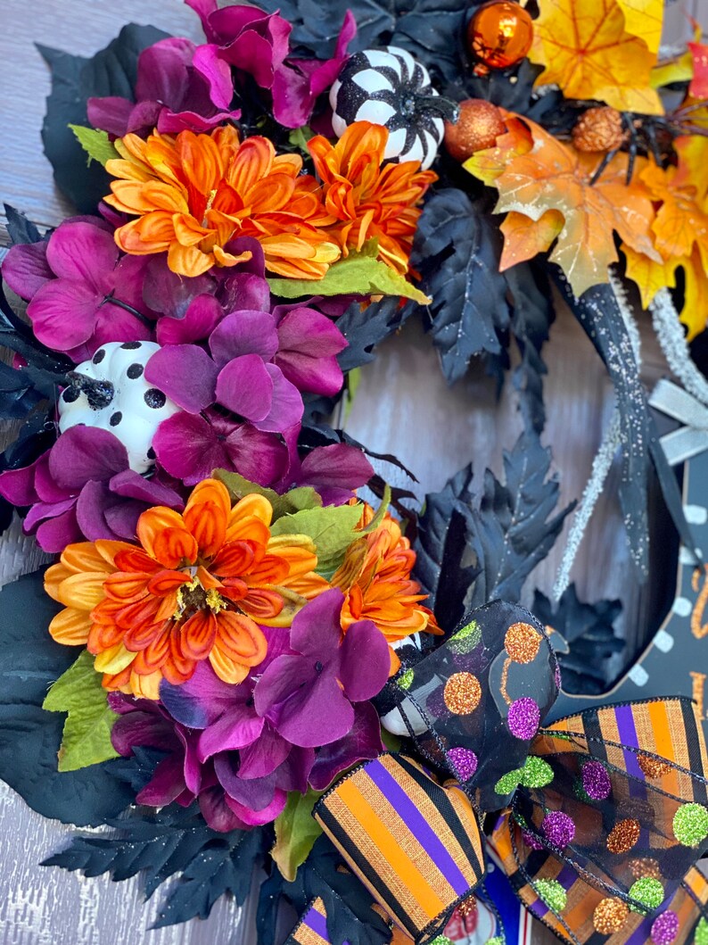 May include: A Halloween wreath with black, orange, and purple flowers, black leaves, and a black and white polka dot pumpkin. The wreath is decorated with a black and orange striped ribbon and a black mesh ribbon with green and orange polka dots.