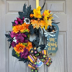 May include: A Halloween wreath with a black witch's boot sign that says "The Witch Is In". The wreath is decorated with fall leaves, black and white polka dot fabric, and purple and orange flowers.