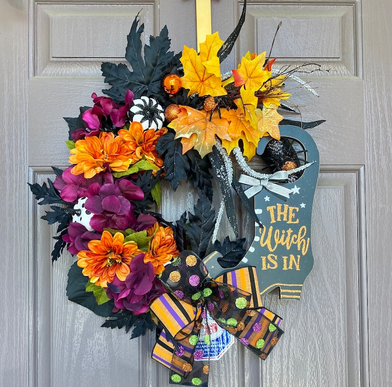 May include: A Halloween wreath with a black witch's boot sign that says "The Witch Is In". The wreath is decorated with fall leaves, black and white polka dot fabric, and purple and orange flowers.