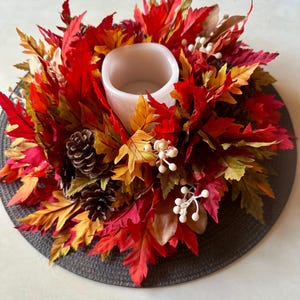 K&ouml;nnte beinhalten: Ein herbstliches Kerzenhalter mit einer wei&szlig;en Kerzenschale, umgeben von leuchtenden roten, orangefarbenen und gelben Kunstbl&auml;ttern. Die Anordnung umfasst Tannenzapfen und wei&szlig;e Beerenakzente, die ein festliches Herbst-Arrangement bilden.