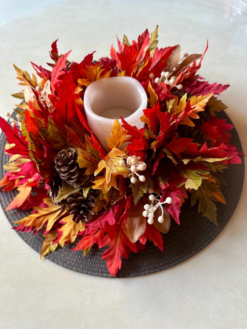 K&ouml;nnte beinhalten: Ein herbstliches Kerzenhalter mit einer wei&szlig;en Kerzenschale, umgeben von leuchtenden roten, orangefarbenen und gelben Kunstbl&auml;ttern. Die Anordnung umfasst Tannenzapfen und wei&szlig;e Beerenakzente, die ein festliches Herbst-Arrangement bilden.