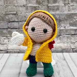 May include: A crocheted doll wearing a yellow raincoat, green boots, and a red flower. The doll has brown hair and is holding a white crocheted object.