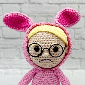 May include: A pink crocheted bunny rabbit doll wearing glasses and pink bunny slippers. The doll has a yellow yarn headband and a crocheted face.