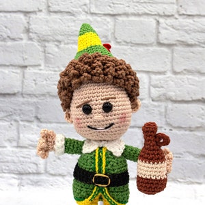 May include: A crocheted elf doll wearing a green and yellow hat, green and yellow pants, and a black belt. The elf is holding a brown crocheted bottle.