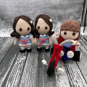 May include: Two crocheted dolls with brown hair and blue dresses stand side-by-side. A third crocheted doll with brown hair is riding a red tricycle.