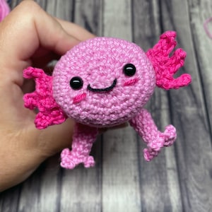 May include: Pink crocheted axolotl plushie with black eyes and a smiling mouth. The axolotl has pink frilly gills and is holding its arms up.
