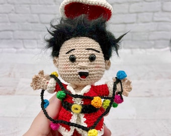 May include: A crocheted Christmas ornament of a smiling boy wearing a Santa hat and a red and white Santa suit with a string of colorful lights wrapped around him.