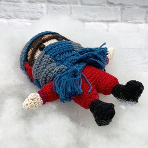May include: A crocheted toy with a red and white striped body, blue scarf, and black boots. The toy has a brown face with black eyes and a brown beard.