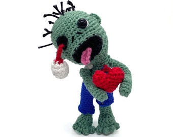 May include: A crocheted zombie plush toy with a green body, blue pants, and a red heart. The zombie has a missing eye and a tongue sticking out.