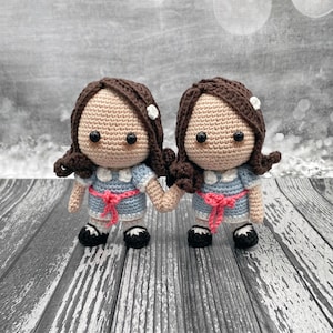 May include: Two crocheted dolls wearing blue dresses with pink sashes and white collars. The dolls have brown hair and black eyes. They are holding hands and standing on a wooden surface.