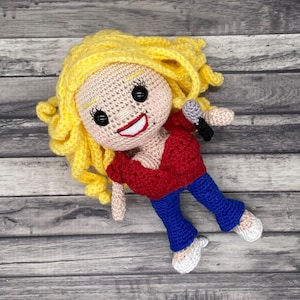 May include: A crocheted doll with blonde hair, a red top, blue jeans, and white shoes. The doll is holding a microphone.