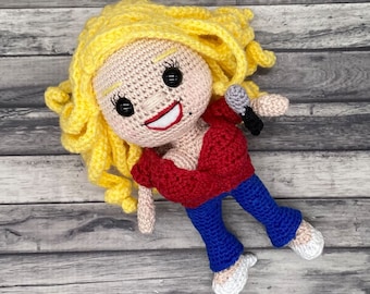 Peut inclure: Une poupée en crochet avec des cheveux blonds, un haut rouge, un jean bleu et des chaussures blanches. La poupée tient un microphone.