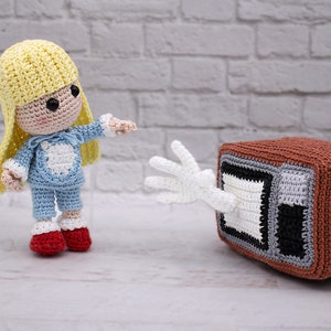 May include: A crocheted doll with blonde hair wearing blue pyjamas reaches out to a crocheted television with a white hand.