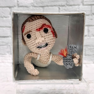 Puede incluir: Una muñeca de crochet de una persona con una raya roja en la frente, sosteniendo una pistola de crochet gris con una llama roja. La muñeca está en una caja de plástico transparente.