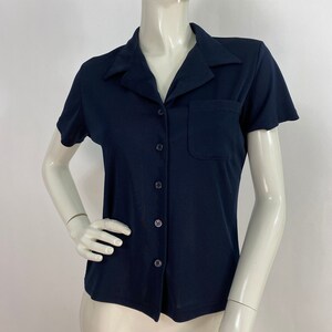 May include: A navy blue short-sleeved shirt with a collared neckline and a single pocket on the chest. The shirt has a button-down front and is displayed on a mannequin. The fabric appears to be a smooth, knit material.