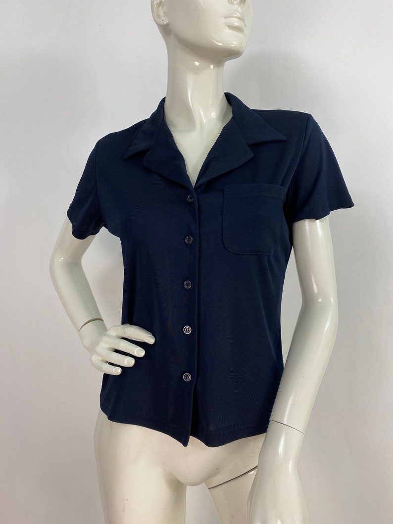 May include: A navy blue short-sleeved shirt with a collared neckline and a single pocket on the chest. The shirt has a button-down front and is displayed on a mannequin. The fabric appears to be a smooth, knit material.