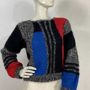 May include: A grey, black, blue and red geometric patterned jumper. The jumper has a crew neck and long sleeves.