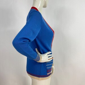 May include: A blue cardigan with red and cream trim. The cardigan has a V-neck and a pattern on the front.