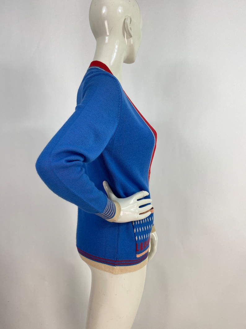 May include: A blue cardigan with red and cream trim. The cardigan has a V-neck and a pattern on the front.