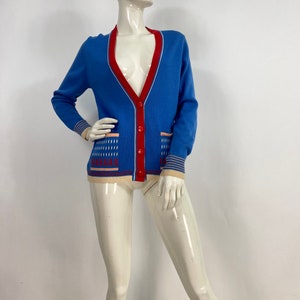 May include: A blue cardigan with a red trim and a white and pink patterned design. The cardigan has a button closure and two pockets.