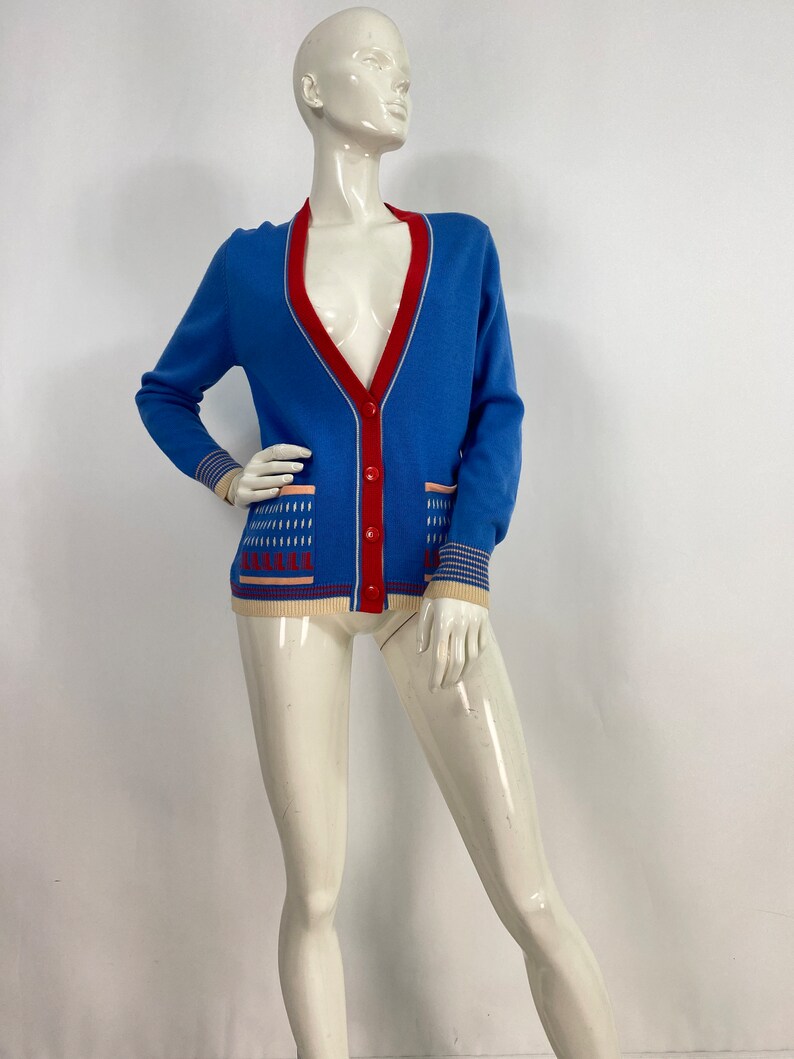 May include: A blue cardigan with a red trim and a white and pink patterned design. The cardigan has a button closure and two pockets.