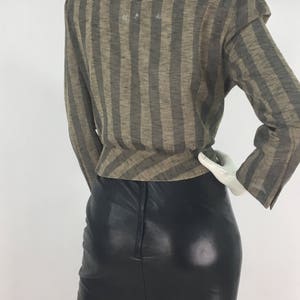 Puede incluir: Una blusa de manga larga con rayas marrones y negras y cuello en V. La blusa se lleva sobre una falda de cuero negra.