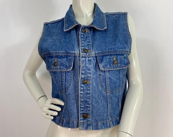 May include: A sleeveless denim vest in a medium wash blue. The vest features a collared neckline, two chest pockets, and brass-colored buttons down the front. The vest is displayed on a mannequin.