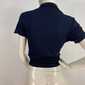 May include: Navy blue short-sleeved polo shirt. The shirt has a collared neckline and a fitted silhouette. The back view shows the shirt's tailored design, emphasizing its shape and style. The fabric appears smooth and has a slight sheen.