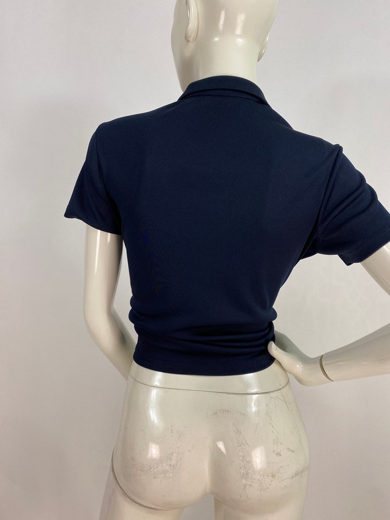May include: Navy blue short-sleeved polo shirt. The shirt has a collared neckline and a fitted silhouette. The back view shows the shirt's tailored design, emphasizing its shape and style. The fabric appears smooth and has a slight sheen.