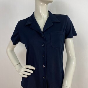 May include: A navy blue short-sleeved button-down shirt with a pocket on the chest. The shirt has a classic collar and is displayed on a mannequin. The shirt is made of a smooth, solid-colored fabric.