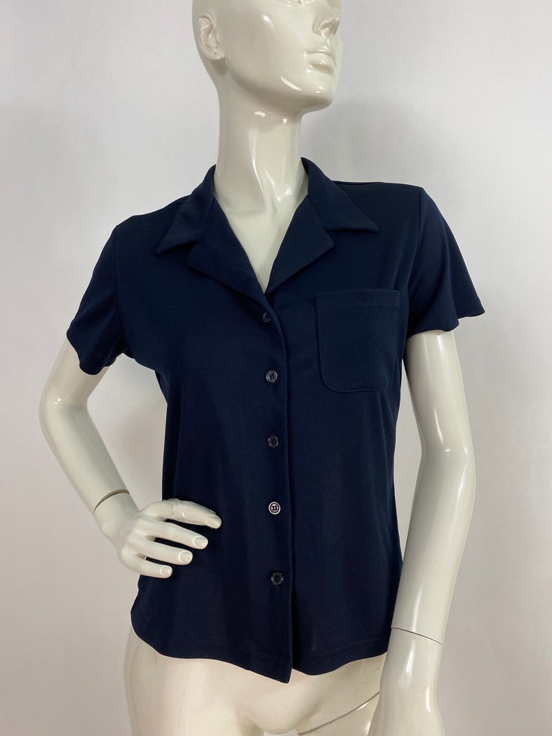 May include: A navy blue short-sleeved button-down shirt with a pocket on the chest. The shirt has a classic collar and is displayed on a mannequin. The shirt is made of a smooth, solid-colored fabric.