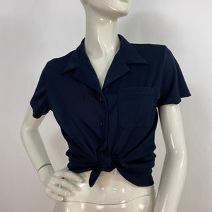 May include: Navy blue short-sleeve shirt with a collar, pocket, and button closure. The shirt is tied at the waist. The shirt is displayed on a mannequin.