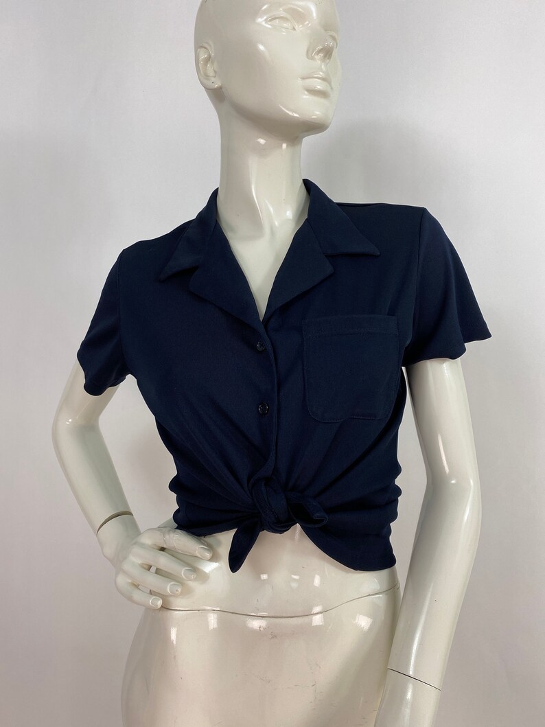 May include: Navy blue short-sleeve shirt with a collar, pocket, and button closure. The shirt is tied at the waist. The shirt is displayed on a mannequin.