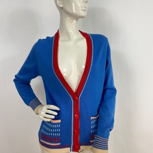 May include: A blue cardigan with a red and white trim, two pockets, and a button closure. The cardigan has a geometric pattern on the bottom.