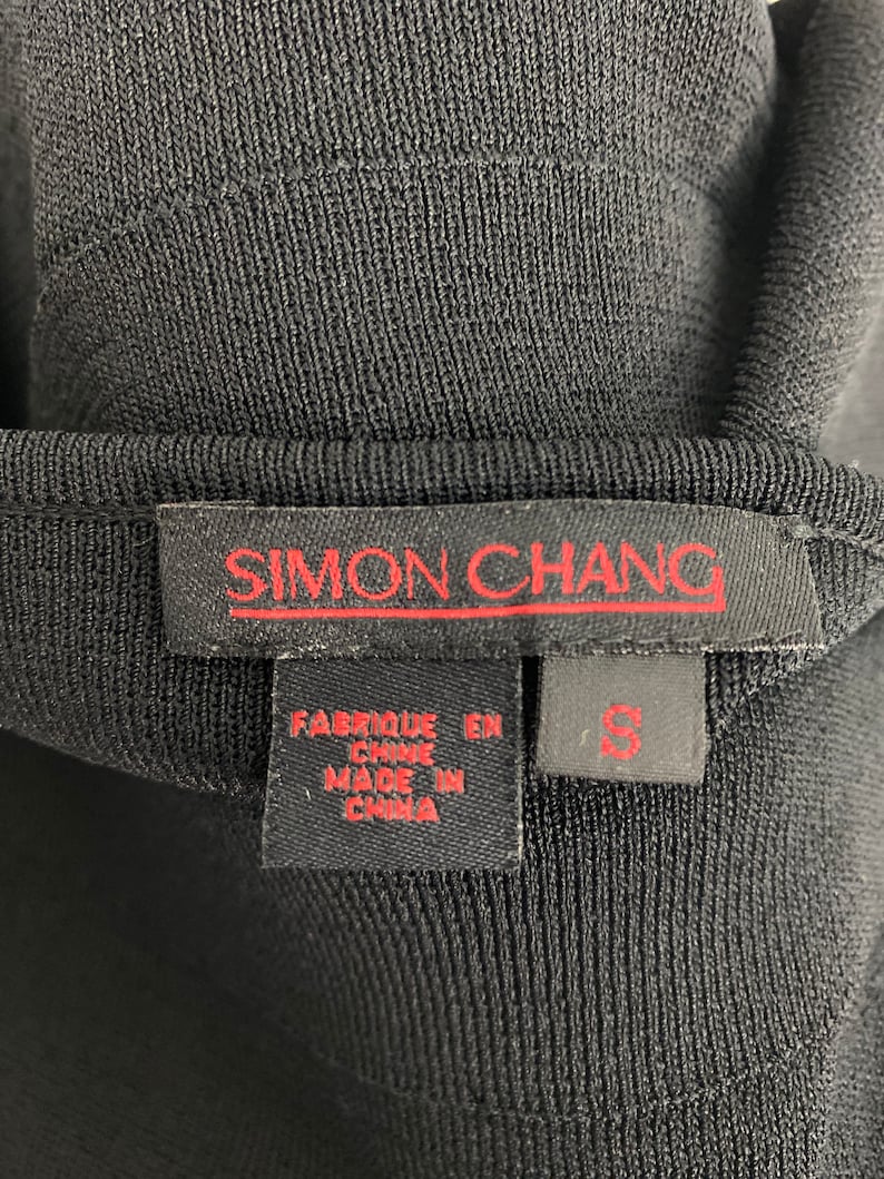 K&ouml;nnte beinhalten: Schwarzes Strickoberteil mit einem roten "SIMON CHANG"-Etikett und einem Gr&ouml;&szlig;enetikett "S". Das Etikett tr&auml;gt au&szlig;erdem die Aufschrift "Fabrique en Chine" und "Made in China".