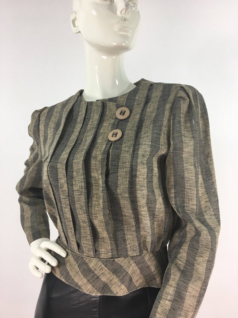 Puede incluir: Una blusa de manga larga a rayas con cintura ajustada y un dobladillo ligeramente acampanado. La blusa tiene un escote redondo y dos botones grandes en la parte delantera.