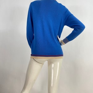 May include: A blue long-sleeved sweater with a white and blue striped cuff and a beige and burgundy trim around the neck and bottom.