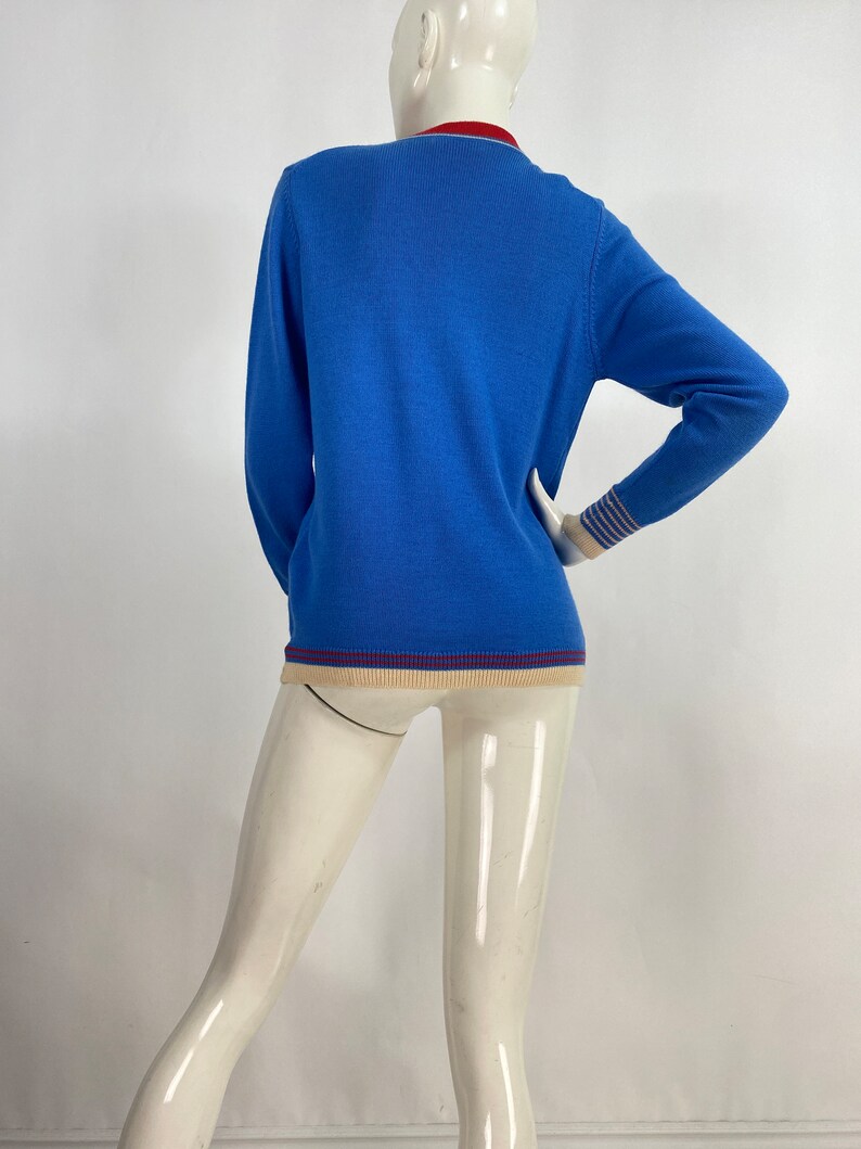 May include: A blue long-sleeved sweater with a white and blue striped cuff and a beige and burgundy trim around the neck and bottom.