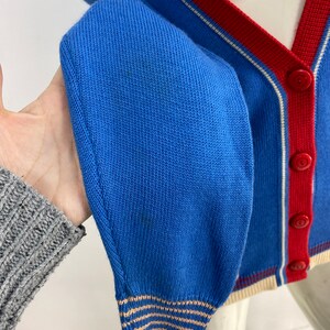 May include: A blue cardigan with a red and white striped trim. The cardigan has a button closure and a ribbed knit texture.