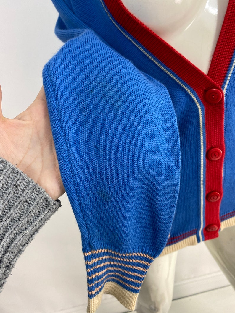 May include: A blue cardigan with a red and white striped trim. The cardigan has a button closure and a ribbed knit texture.