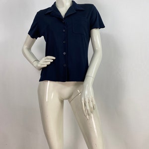 May include: A navy blue short-sleeved shirt with a collared neckline and a single chest pocket. The shirt has a button-down front and is displayed on a mannequin. The fabric appears to be a smooth, solid color.