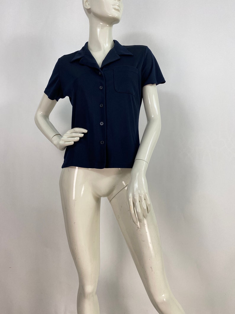 May include: A navy blue short-sleeved shirt with a collared neckline and a single chest pocket. The shirt has a button-down front and is displayed on a mannequin. The fabric appears to be a smooth, solid color.