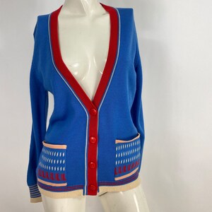 May include: A blue cardigan with a red and white trim. The cardigan has two pockets with a geometric pattern in white, red, and pink.