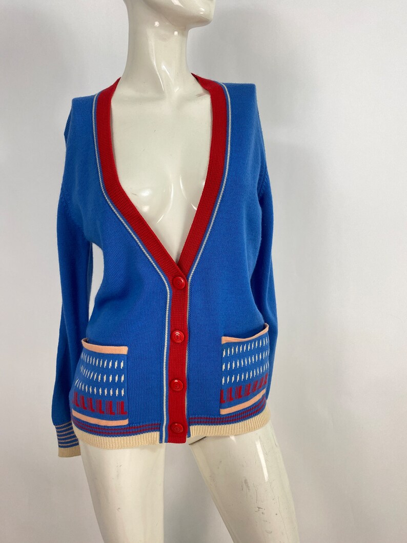 May include: A blue cardigan with a red and white trim. The cardigan has two pockets with a geometric pattern in white, red, and pink.