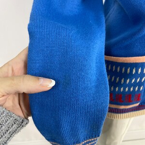 May include: Close-up of the blue knitted cuff of a sweater with a white and pink striped trim. The sweater has a pattern of white and pink stripes and a small white design.