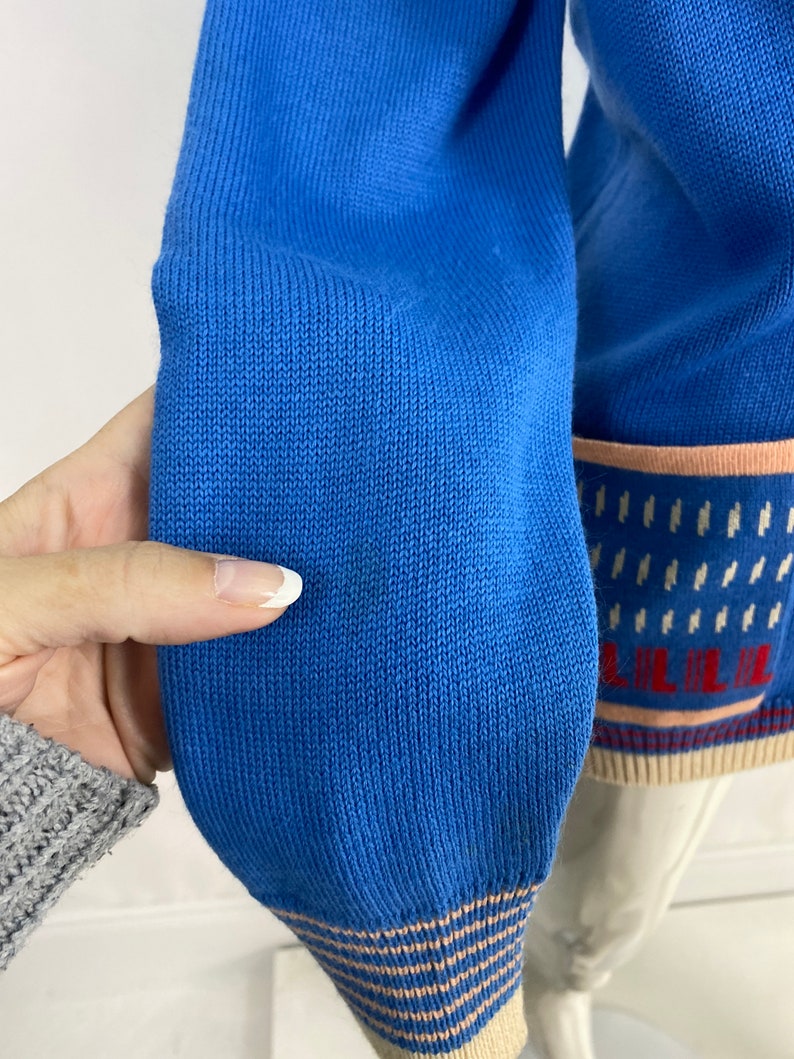 May include: Close-up of the blue knitted cuff of a sweater with a white and pink striped trim. The sweater has a pattern of white and pink stripes and a small white design.