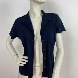 May include: A navy blue short-sleeved shirt with a collared neckline and a single chest pocket. The shirt has a button-up front and is displayed on a mannequin. The fabric appears to be a lightweight knit.
